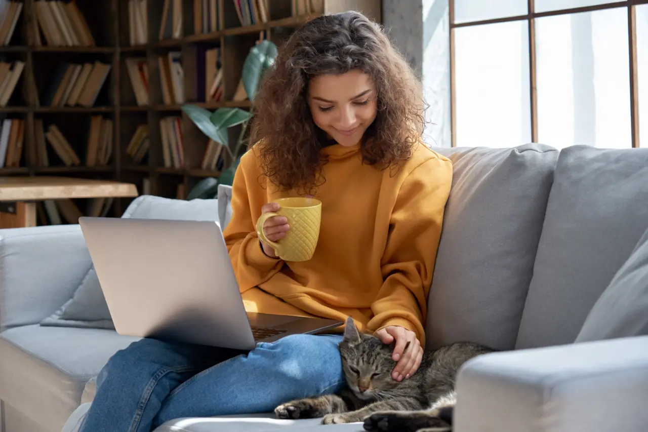 Hispanic girl holding a yellow mug and sitting on the couch with a laptop on her lap while petting her cat next to her