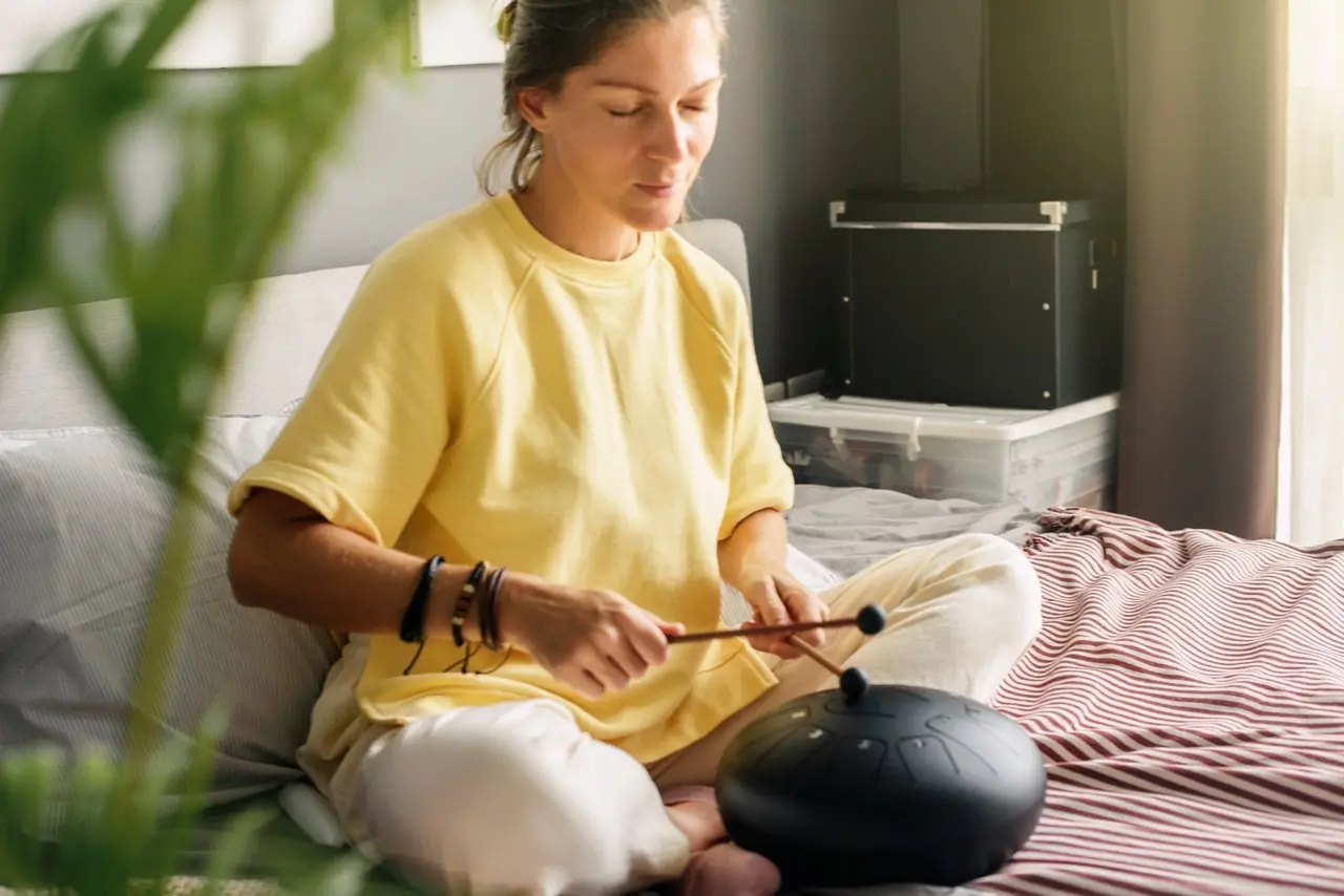 Holistic Therapy: woman in a yellow shirt sitting cross-legged on her bed with her eyes closed playing a Steel Tongue Drum