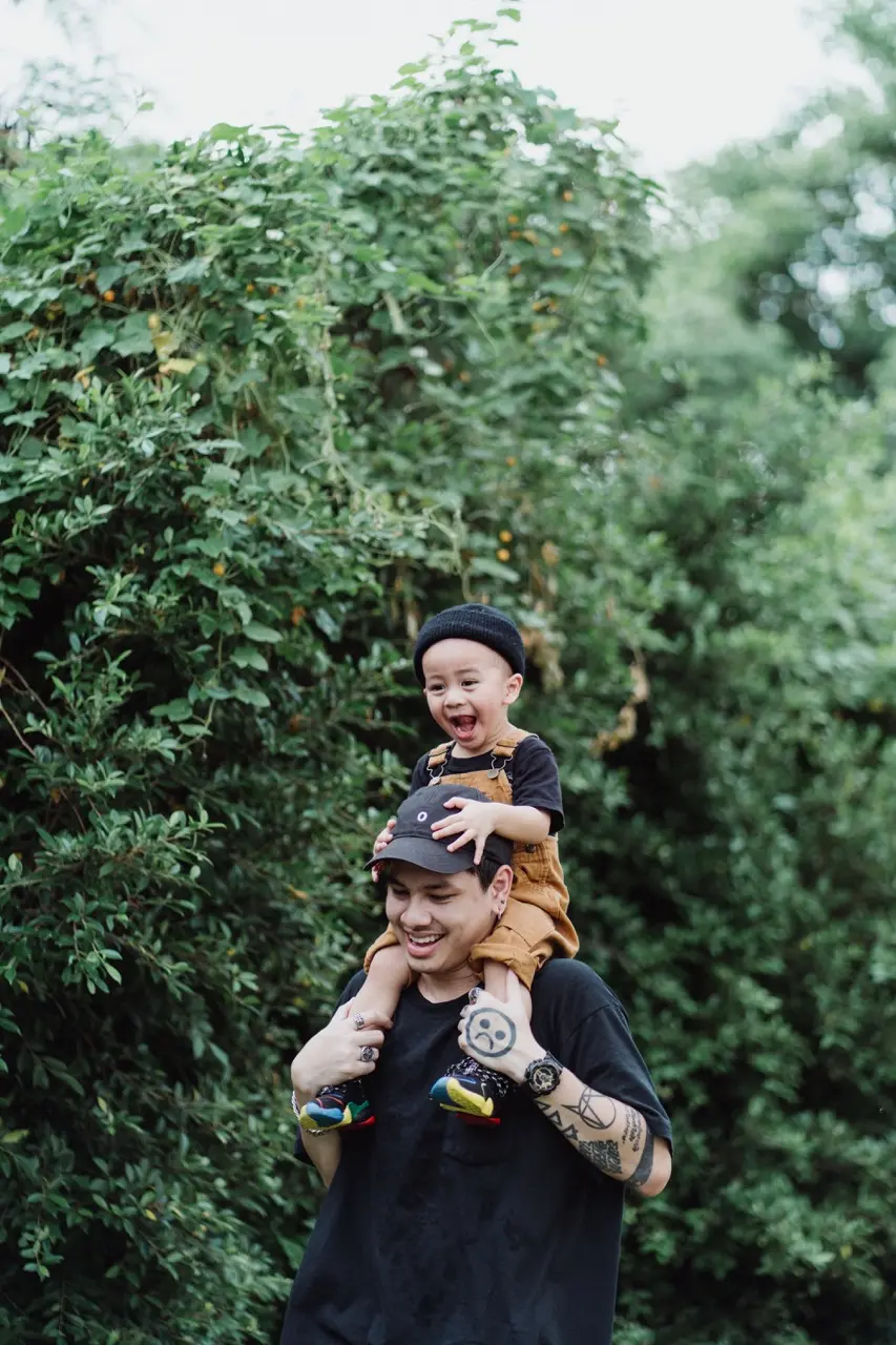 A man taking a walk outside with his son sitting on his shoulders, both smiling and laughing